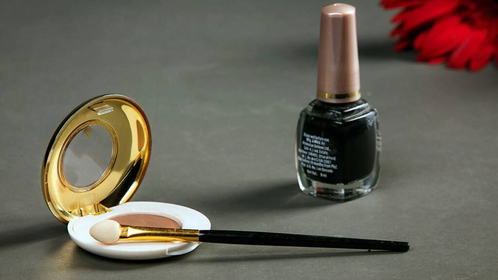 An open compact with a mirror and a sponge applicator is on a gray surface. Next to it, there is a bottle of dark nail polish with a beige cap. A red flower is out of focus in the top right corner. The background is a plain dark gray.