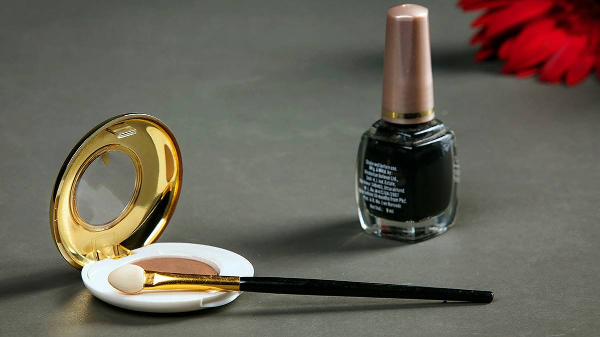 An open compact with a mirror and a sponge applicator is on a gray surface. Next to it, there is a bottle of dark nail polish with a beige cap. A red flower is out of focus in the top right corner. The background is a plain dark gray.